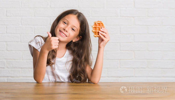 年轻的西班牙裔小孩坐在桌子上吃华夫饼，开心地大笑着做，好的，用手指签个名，好的