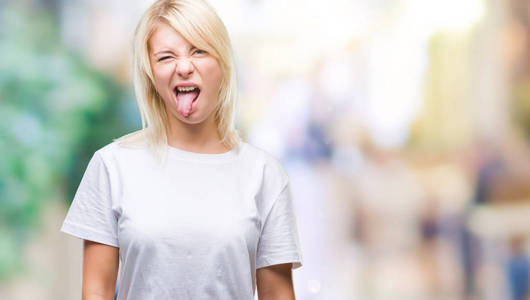 年轻漂亮的金发女人穿着白色T恤，在孤立的背景上伸出舌头，以有趣的表情高兴。 情感概念。
