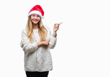 年轻漂亮的金发女人戴着圣诞帽，在孤立的背景上，脸上挂着一个大大的微笑，用手和手指指向一边，看着相机。