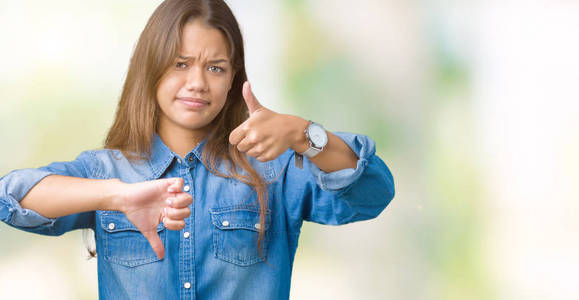 年轻漂亮的黑发女人穿着蓝色牛仔衬衫，在孤立的背景下做拇指上下的分歧和一致的表达。 疯狂冲突