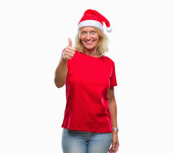 中年金发女人戴着圣诞帽，戴着孤立的背景，用手做快乐的竖起大拇指的手势。 赞许的表情看着相机，显示出成功。