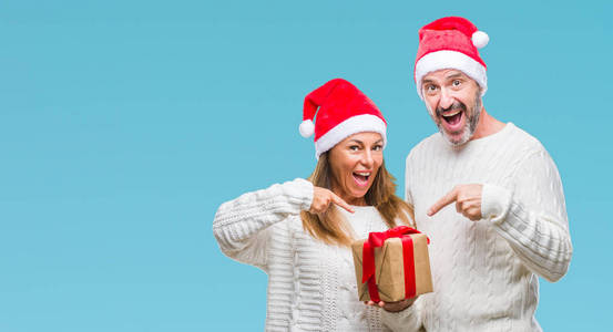 中年西班牙夫妇戴着圣诞帽，在孤立的背景上拿着礼物，非常高兴地用手和手指指着。