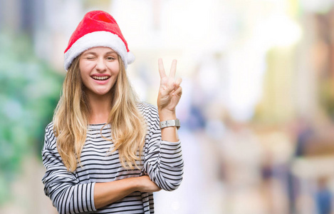年轻漂亮的金发女人戴着圣诞帽，在孤立的背景上微笑，快乐的脸对着镜头眨着眼睛，做着胜利的标志。 二号。