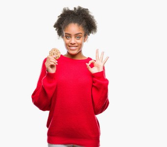 年轻的非洲美国女人吃巧克力饼干，在孤立的背景下做OK标志，手指，优秀的象征