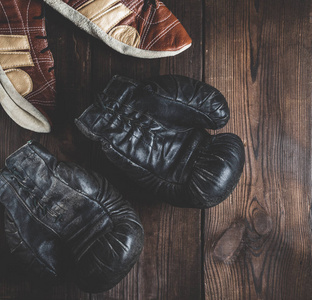 一双带鞋带的非常旧的皮革黑色拳击手套和一双棕色木制背景的皮鞋，复古色调