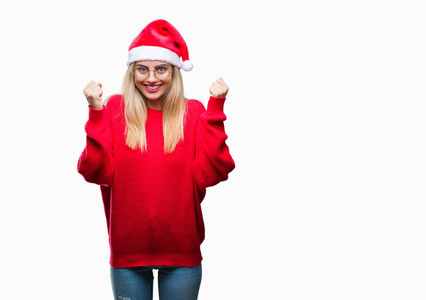 年轻漂亮的金发女人戴着圣诞帽，在孤立的背景上庆祝，惊讶和惊讶的成功，举起手臂和睁开眼睛。 赢家概念。