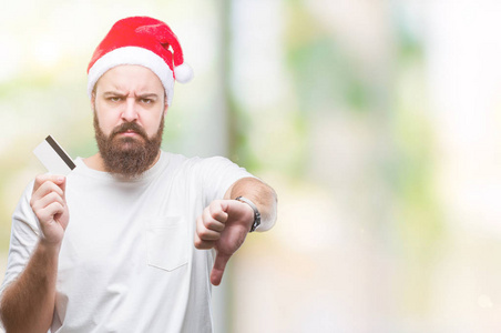 年轻的白种人戴着圣诞帽，在孤立的背景下拿着信用卡，愤怒的脸，消极的迹象，表示不喜欢，竖起大拇指拒绝概念