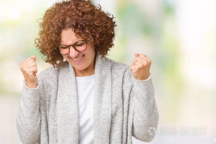 漂亮的中老年妇女穿着夹克和眼镜，在孤立的背景下，非常高兴和兴奋地做着胜利者的手势，手臂抬起，微笑着，尖叫着，争取成功。 庆祝概念