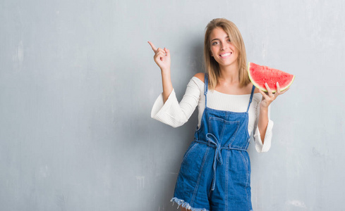 美丽的年轻女子在灰墙上吃西瓜，非常高兴地用手和手指指向一边