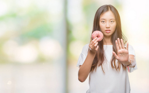 年轻的亚洲女人在孤立的背景下吃甜甜圈，张开手做停止标志，严肃自信的表情防御手势