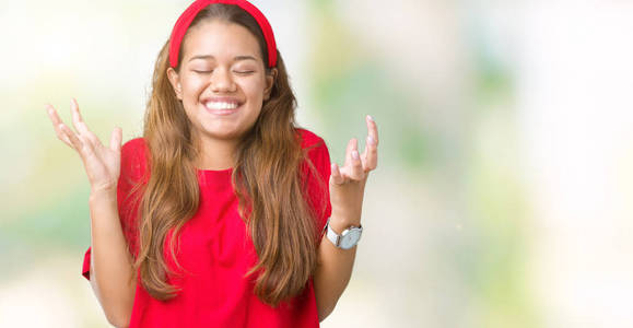 年轻漂亮的黑发女人穿着红色T恤在孤立的背景上庆祝疯狂和疯狂的成功，手臂抬起和闭上的眼睛尖叫兴奋。 赢家概念
