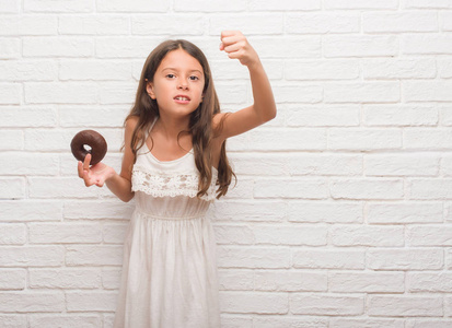 年轻的西班牙裔小孩在白砖墙上吃巧克力甜甜圈，生气和沮丧，愤怒，疯狂地大喊大叫，举起手愤怒的概念