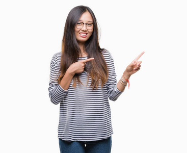 年轻的亚洲女人戴着眼镜，在孤立的背景上微笑，看着相机，双手和手指指向一边。
