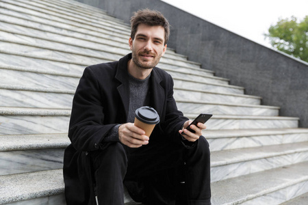 英俊的年轻商人坐在户外用手机喝咖啡的形象。