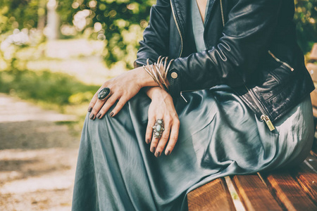 时尚的细节，一个女人穿着丝绸连衣裙，皮夹克和戒指和手镯作为配饰，坐在公园的长凳上的特写