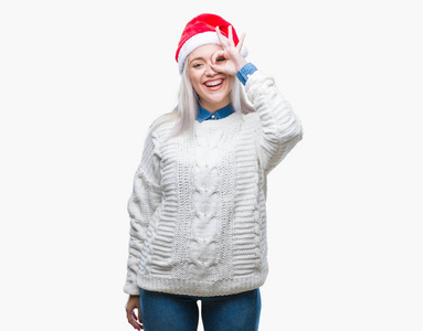 年轻的金发女人戴着圣诞帽，在孤立的背景上做着好的手势，用手微笑的眼睛透过手指看着幸福的脸。