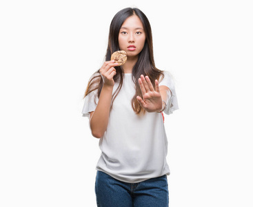 年轻的亚洲妇女吃巧克力片饼干孤立的背景和开放的手做停止标志，严肃和自信的表情防御手势