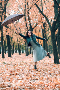 带着雨伞在秋天公园摆姿势的女人。 明亮的黄色叶子和树木。 她模仿风。