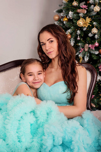 女孩抱着妈妈坐在一棵新年树旁，穿着郁郁葱葱的蓝色晚礼服