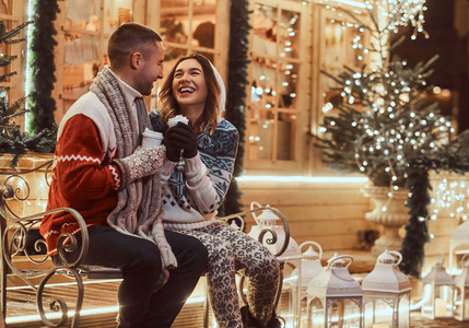 一对年轻的浪漫夫妇在圣诞节期间穿着温暖的衣服在户外, 坐在长凳上, 在装饰着美丽灯光的晚间街道上, 用热咖啡取暖