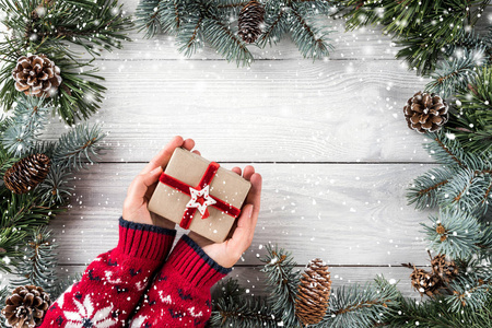 女性手拿着圣诞礼品盒在白色的木制背景上，有杉树枝松果雪花。 圣诞节和新年快乐主题。 平躺式顶部视图