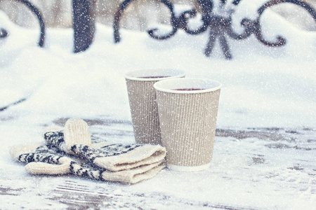 两个纸制酒杯，一杯热饮料，咖啡，可可，覆盖葡萄酒和白色和黑色手套在户外的桌子上在冬天。冬季时间概念。在寒冷中晒太阳。