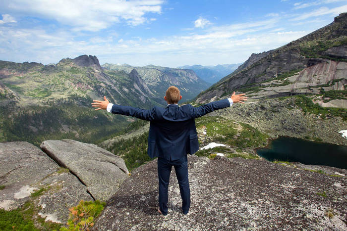 一位身穿商务服的年轻人在阳光明媚的夏日在群山和蓝天的背景下在山顶
