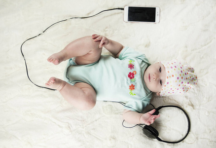 明亮的小美丽的新生婴儿躺在床上听音乐，旁边的耳机和电话关闭