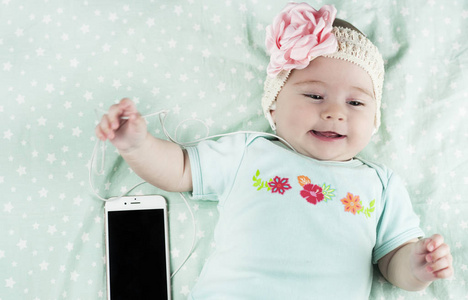 明亮的小美丽的新生婴儿躺在床上听音乐，旁边的耳机和电话关闭