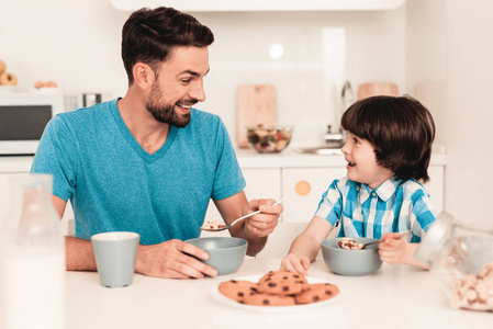 微笑的儿子和父亲在厨房吃早餐。 穿衬衫的男孩。 现代厨房。 坐着的男孩。 有勺子的男孩。 早上吃早餐。 厨房里的白色桌子。 桌子