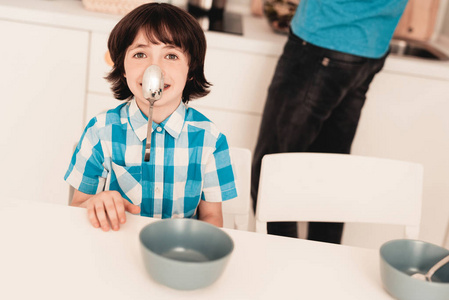 穿衬衫的小男孩早上在厨房玩得很开心。 儿子和父亲。 现代厨房。 坐着的男孩。 有勺子的男孩。 早上吃早餐。 厨房里的白色桌子。 