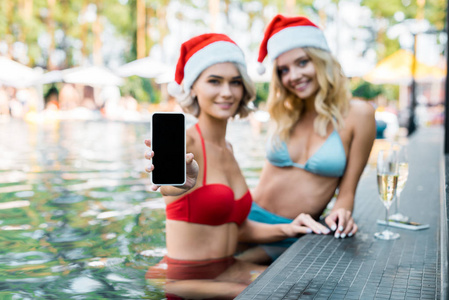 穿着泳衣和圣诞老人帽的微笑女性，在游泳池放松时展示带有空白屏幕的智能手机