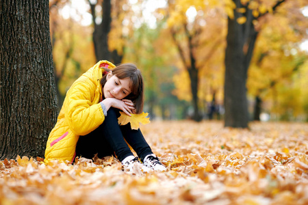 悲伤的少女坐在秋天公园的树旁。 明亮的黄色叶子和树木。