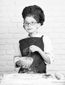 红色制服的小男孩可爱厨师厨师和帽子上戴着眼镜，站在旁边用擀面杖表和烹饪，还有厨房沾满泪水的脸面粉搅拌在白砖的背景墙上的绿松石碗