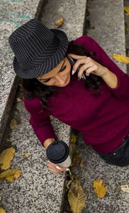 一位穿着红色毛衣带纸杯的年轻女士坐在满是落叶的楼梯上，一边打电话，一边高角度观看