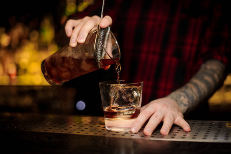 酒保把新鲜的烈性威士忌鸡尾酒倒入一个杯子里，酒吧上有一个冰块。
