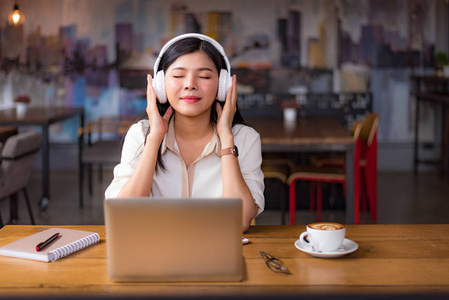 美丽的亚洲女人在咖啡馆里放松和听音乐，有笔记本电脑和咖啡杯。 人和生活方式的概念。 自由职业快乐工作场所主题。 大学和学院的主题