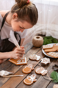 一个年轻女孩在冬天的早晨装饰姜饼。女人在蜂蜜姜饼房子里画冰。木棕色的桌子。复制空间。空白饼干姜饼屋, 准备装修