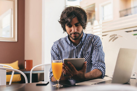 年轻的帅哥在咖啡馆拿着平板电脑，用他的笔记本电脑在线工作，喝着橙汁，轻松自信。