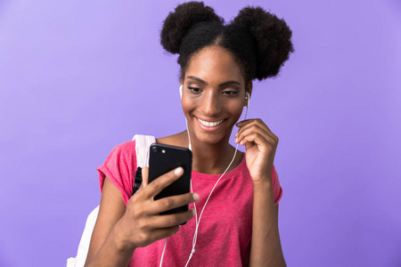 内容照片非裔美国女学生戴背包和白色耳机，手持智能手机隔离在紫罗兰背景上