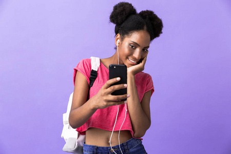 照片积极的非裔美国女学生戴着背包和白色耳机，手持智能手机隔离在紫罗兰背景上