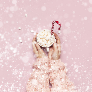 圣诞新年饮料，白色杯子与棉花糖在女性手中针织毛衣和糖果罐在粉红色背景顶部视图平躺冬季传统食物。节日装饰白雪效果