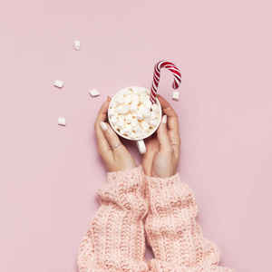圣诞新年饮料，白色杯子与棉花糖在女性手中针织毛衣和糖果罐在粉红色背景顶部视图平躺冬季传统食物。节日装饰庆祝圣诞