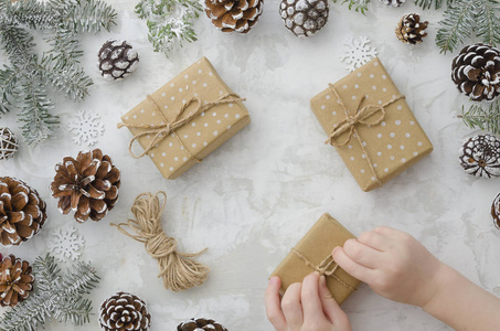 孩子正在用自己的双手包装礼物。顶部视图。平放置礼品盒包裹在牛皮纸, 锥, 绳子, 冷杉树枝和雪花在白色的背景。冬天