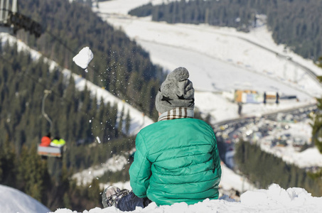 男孩坐在山上的雪坡上，在缆车针叶林和山脉的背景下扔雪球。滑雪胜地。冬季