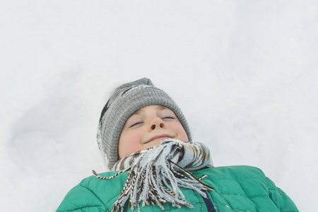 戴着帽子围巾和绿色夹克的男孩躺在雪地里，闭着眼睛。肖像。特写镜头。冬季