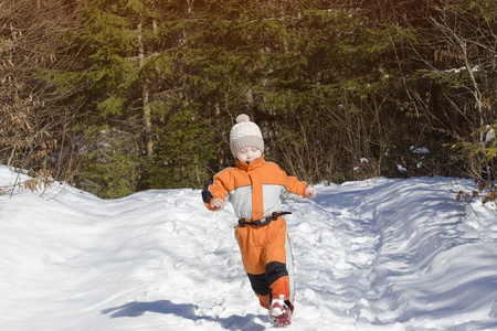 穿着橙色连衣裙的小男孩在一片针叶林的白雪覆盖的道路上奔跑。冬季阳光明媚