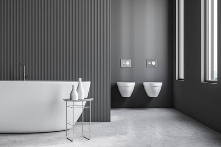 现代浴室的内部有灰色墙壁,石头地板,阁楼窗户,白色浴缸和两个厕所.