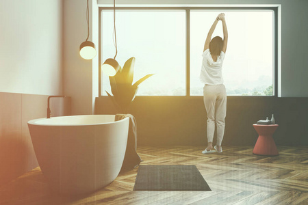 女人在极简主义的浴室内部，白色和灰色的墙壁，木地板，大窗户，白色浴缸与灰色毛巾和粉红色的椅子。盆栽植物。彩色图像双曝光