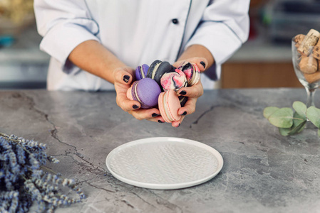 s female hands pour colored macarons into a white plate on a mar